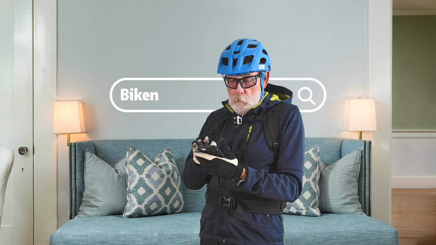 Ein älterer Mann mit Velohelm steht in seinem Wohnzimmer und schaut auf sein Handy